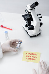 cropped view of scientist holding coronavirus vaccine near microscope and card with covid-19 omicron variant lettering.