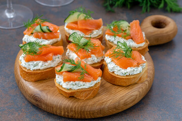 Smoked salmon bruschetta with cream cheese, cucumber and dill served on round wooden board. Close up, selective focus.