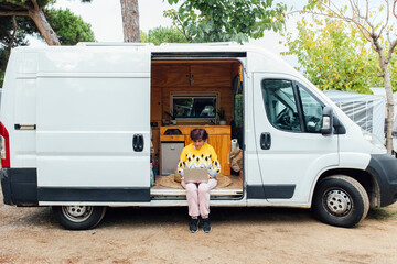 Middle-aged woman sitting with laptop in camper van while working. Nomad life. © EGHStock
