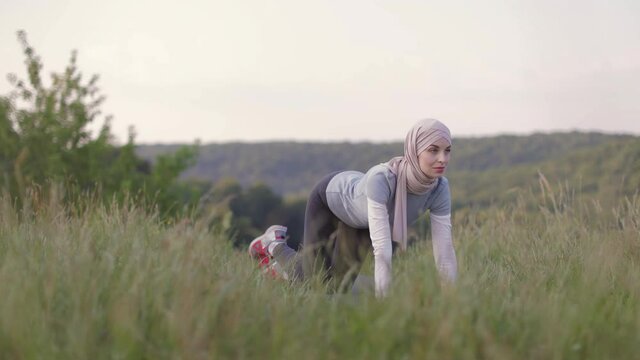 Smiling young woman in hijab and sport outfit doing flexible exercises on yoga mat during morning workout on fresh air. Training at summer park. Healthy lifestyles.