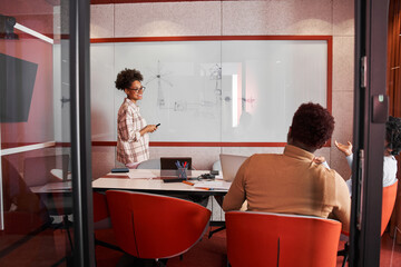 Young multiracial woman standing in front of partners and writing on whiteboard