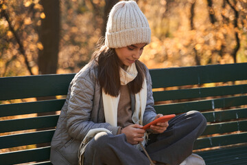 Teenage girl sitting at the bench at the street and turning songs at her smartphone
