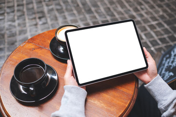 Top view mockup image of a woman holding digital tablet with blank white desktop screen with coffee cups on the table