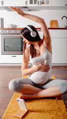 Naklejka premium Pregnant doing workout on orange fitness mat. Pragnant yoga at home in the kitchen. Pregnant in white headphones.