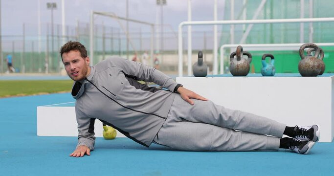 Planking fitness athlete man doing moving side plank exercises or crunches at outdoor gym training obliques muscles on floor. Bodyweight workout outside at athletics stadium. Man in sweatsuit