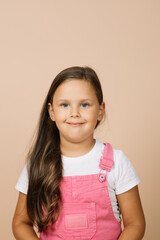 Close up face kid with bright shining eyes and calm happy wide smile looking at camera wearing bright, pink jumpsuit and white t-shirt on beige background.