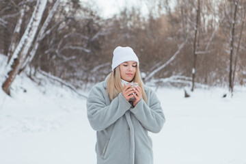 A beautiful young girl smiling and enjoying drinking coffee in a winter forest.