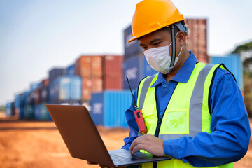 An industrial worker wearing a yellow protective cap holds a laptop to inspect the container yard.