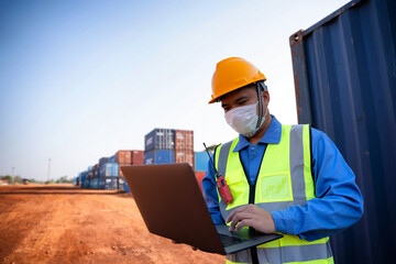 An industrial worker wearing a yellow protective cap holds a laptop to inspect the container yard.