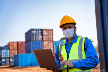 An industrial worker wearing a yellow protective cap holds a laptop to inspect the container yard.