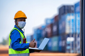 An industrial worker wearing a yellow protective cap holds a laptop to inspect the container yard.