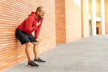 Sportsman wearing a red jacket leaning against a brick wall of clutching his chest acute pain possible heart attack. Hands male holding chest with symptom heart attack disease on city street.
