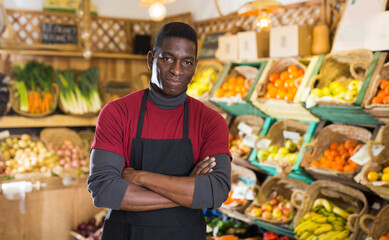 Smiling confident african american greengrocery owner wearing in black apron standing near shelves with fruits and vegetables