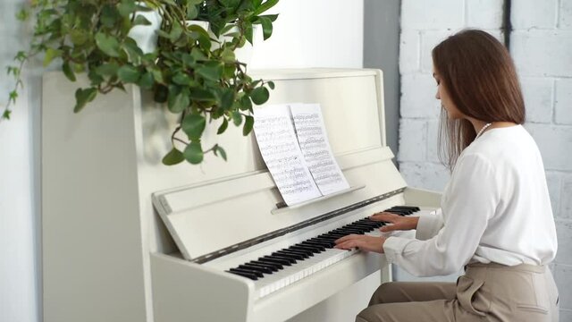 Tracking shot of focused young female musician playing on white classical piano energetic music at home studio during lesson. Lady practicing piano lesson looking on musical notes in classroom.