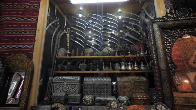 View from left to right of the wall of an antique shop decorated showcasing with variety of silver knives, wooden frame mirrors and filligree pattern trunk boxes.
