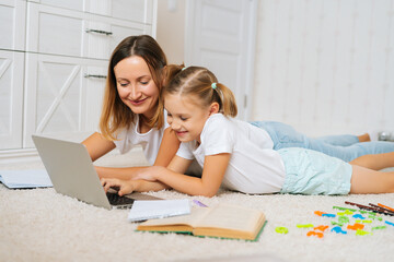 Happy family learning from home together lying on floor in children room looking on laptop screen. Young primary little girl kid studying education with mother. Woman and child laughing feeling fun.