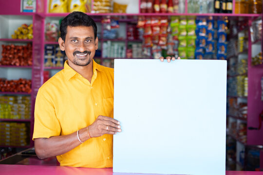 Kirana or groceries Merchant or shopkeeper with Empty sign borad looking at camera - concept of advertisement, promotions and offers on retail store