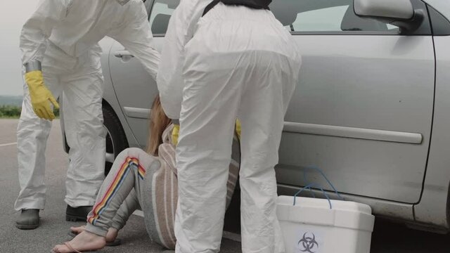 Doctors in bacteriological protection suits caring sick woman sitting next to car outdoors