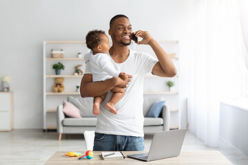 Black Man With Baby Talking On Cellphone While Working At Home Office