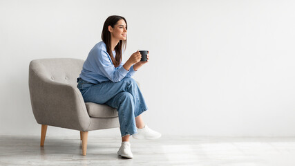 Full length of young woman drinking hot coffee in cozy armchair against white studio wall, banner...
