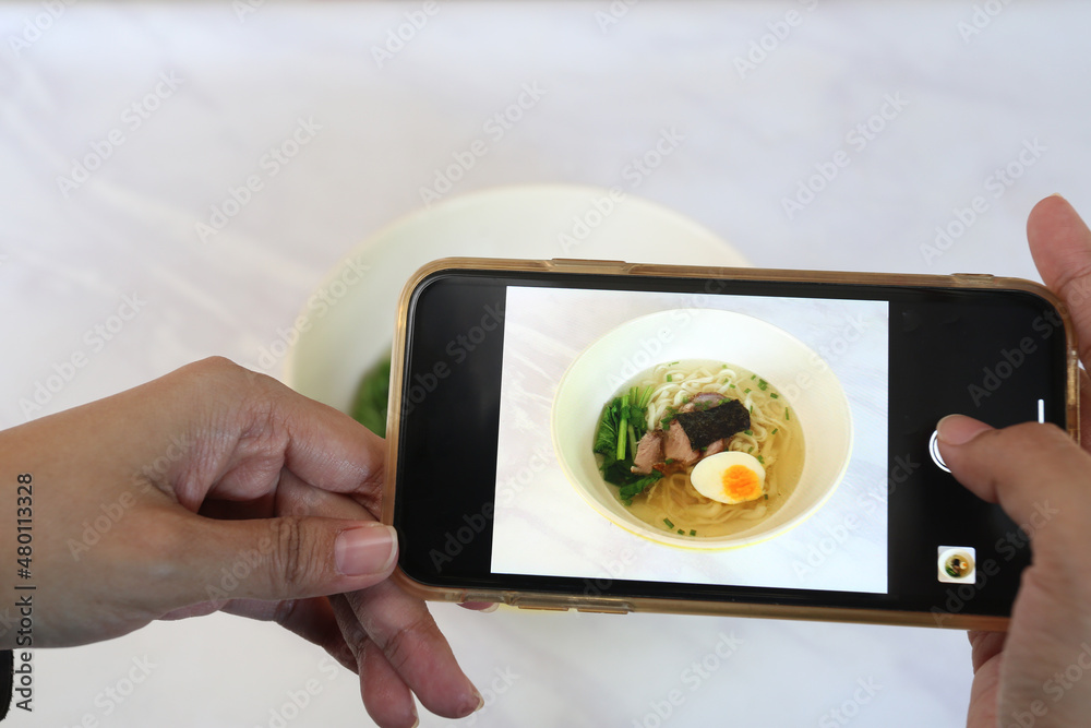 Wall mural Female Hands with the phone close-up pictures of food. The food blogger takes photos of Japanese Ramen food.