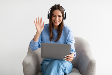 Pretty young lady in headphones waving at laptop screen, sitting in armchair, making video call, chatting online