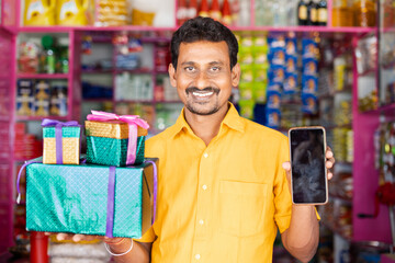 Kirana Shop Merchant with Gifts boxs and mobile phone in hand loking at camera - concpet of Festival sales, promotional offers to win prizes from smartphone.