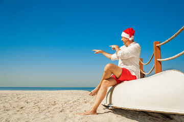 Christmas Santa Claus resting on at ocean sandy tropical beach sitting on a white boat - xmas travel vacation in hot countries concept