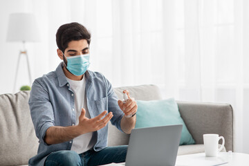 Young arab man in protective face mask disinfecting hands with mist spray while working on laptop at home