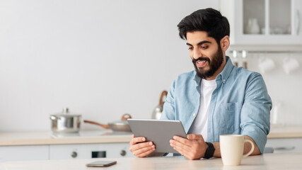 Cheerful arab man using digital tablet while drinking coffee in kitchen at home and checking social networks, copy space