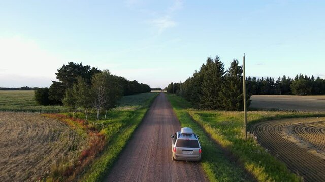 Drone flying fast along a dry and dusty dirt road in rural Alberta during sunset then passing a silver minivan parked on the side of the road.