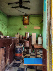 Inside view of Old Indian dirty abandoned hotel kitchen, gods photo and clock hanging on pistachio color painted wall. Gas cylinder, old utensils, wooden table , chair and other objects in the kitchen