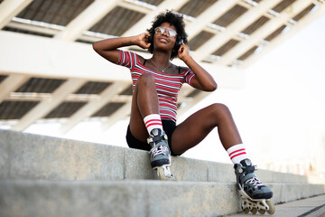 Beautiful African woman with roller skates. Urban sexy girl sitting on the stairs listening the...