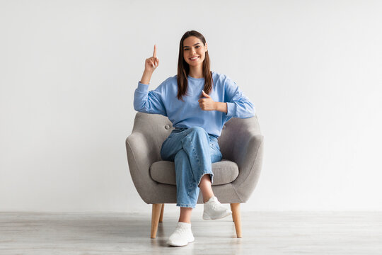 Joyful Young Lady Pointing Upwards And Showing Thumb Up Gesture, Sitting In Armchair Against White Studio Wall