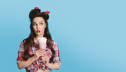 Portrait of sexy pinup woman in retro outfit drinking milk shake looking aside at blank space over blue background