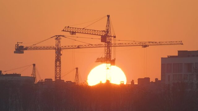 Dark silhouette of tower cranes with big setting sun at high residential apartment buildings construction site at sunset. Real estate development