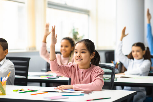 Diverse Group Of Little School Children Raising Hands At Classroom