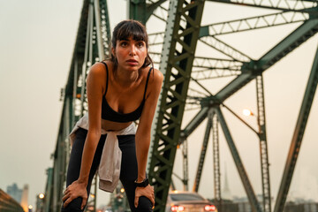 Obraz na płótnie Canvas Confidence Hispanic woman in sportswear jogging on the bridge in the city at summer sunset. Healthy wellness female athlete enjoy outdoor lifestyle sport training workout fitness exercise and running