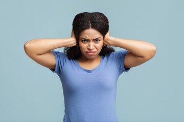 Upset african american woman closing her ears with hands, protecting from loud noise on blue studio background