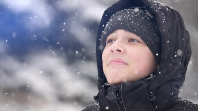 A European boy stands on the street in a heavy snowfall, he looks at the snow and smiles. Close-up, slow motion, shallow depth of field