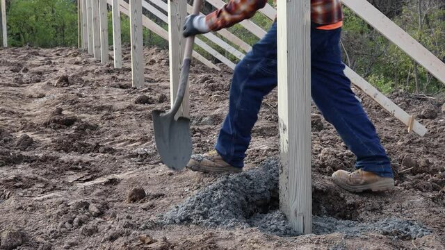 Construction worker using shovel to put cement into post hole for principal pole of new barn construction; uses his foot to level and tamp cement; concepts of new construction and blue collar labor