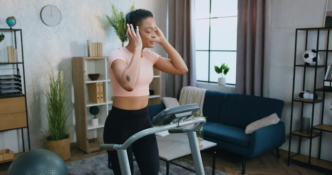 Lovely smiling sporty young african american woman in training clothes running on treadmill and simultaneously listening favourite music in earphones at home