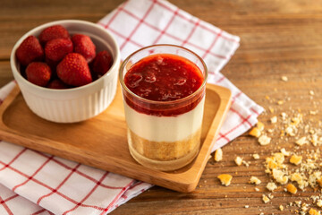 Close up of triple crispy strawberry cheesecake in shot glass on square wooden tray over red, white plaid napkin with some crackers crumble on the table. Eatable recipe decoration dessert concept