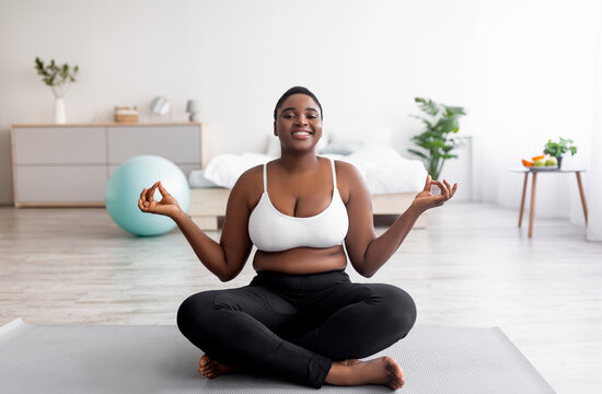 Overweight black woman sitting on sports mat in lotus pose, meditating, doing yoga practice at home