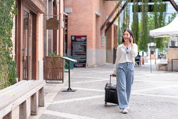 Smiling woman traveller talking using smartphone and dragging black suitcase luggage bag walking to passenger boarding in airport, Travel concept