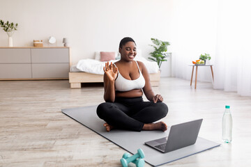 Overweight black woman sitting on yoga mat in front of laptop, greeting personal trainer, waving at...