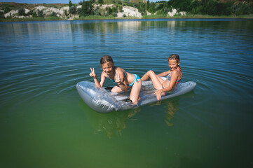 Two little cute girls in swimsuits swim on an inflatable mattress on a blue career lake.