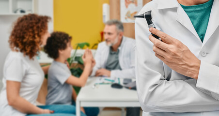 Audiologist with otoscope over background consultation doctor with child patient and his mother at hearing clinic. Ear treatment and hear exam