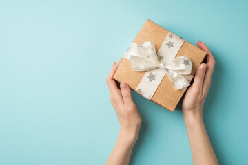 First person top view photo of valentine's day decorations young person's hands demonstrating kraft paper giftbox with white ribbon bow star pattern on isolated pastel blue background with copyspace