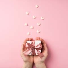 First person vertical top view photo of valentine's day decorations woman's hands holding small white giftbox with pink ribbon bow on palms and small pink hearts on isolated pastel pink background